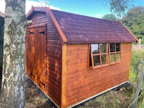 Reddish-brown Dutch Barn shed with gambrel roof, double doors and multi-pane side windows by stone wall