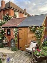 Two interconnected garden sheds – brick section with tiled roof and wooden shed with felt roof – in a cottage garden with climbing roses and clematis