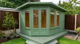 The Haven Corner Summerhouse in sage green with double doors and full-height glazing on matching green decking in a garden corner
