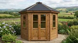 The Panoramic Octagon Summerhouse with timber cladding, dark shingle roof and double glazed doors on flagstone patio in countryside garden
