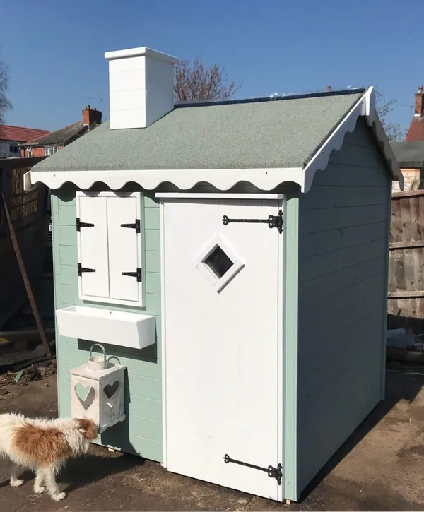 Pastel cottage playhouse with white chimney, shutters, planter box and scalloped roof trim