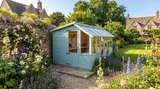 Light blue potting shed with integrated greenhouse section in a cottage garden with lavender and hollyhocks