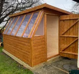 Exterior view of a timber potting shed with a large slanted window front and side door open, showing the spacious interior