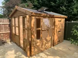 Exterior view of a large timber potting shed featuring extensive floor-to-ceiling glazing on two sides and a separate integrated storage section with a solid door