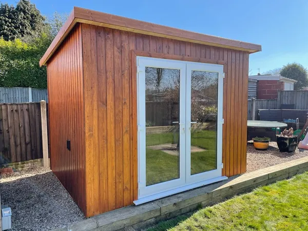 The Modern Pent Garden Room with vertical brown timber cladding and white French doors in a sunny garden